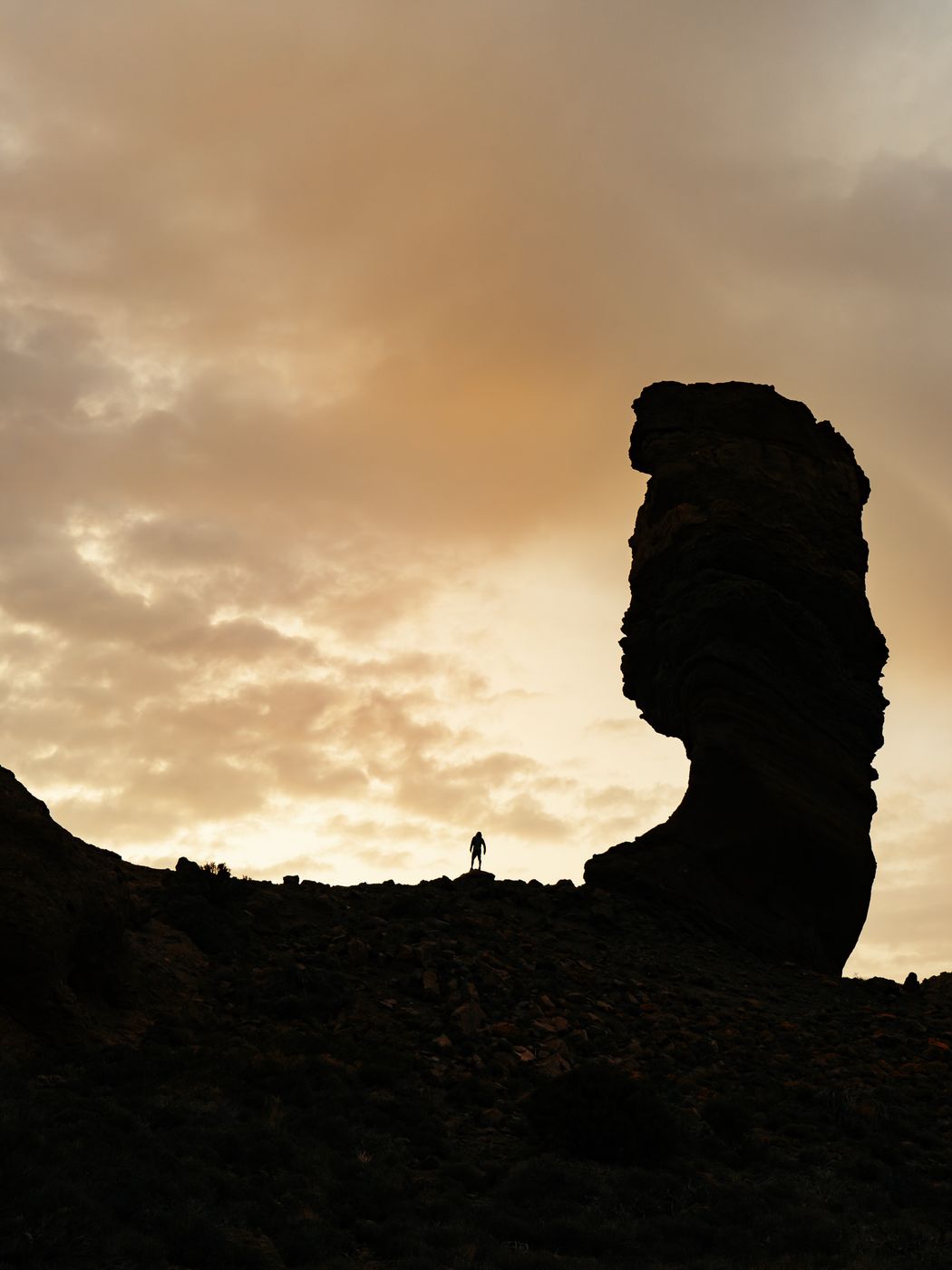 Hiker silhouetted below a tall rock column at sunset
